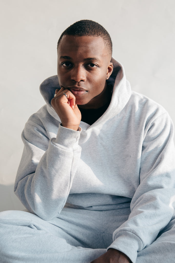 A young Black man in a gray hooded sweatshirt posing with his hand on his chin posing for the cover image of A Real One - the new fashion company launched by Shaun King designed to shake up the supply chain to be Black-owned from farm to closet.  